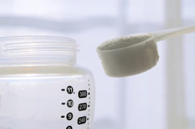 A close-up of a baby bottle with measurement markings and a filled scoop of powdered formula above the bottle opening, ready to be poured in.