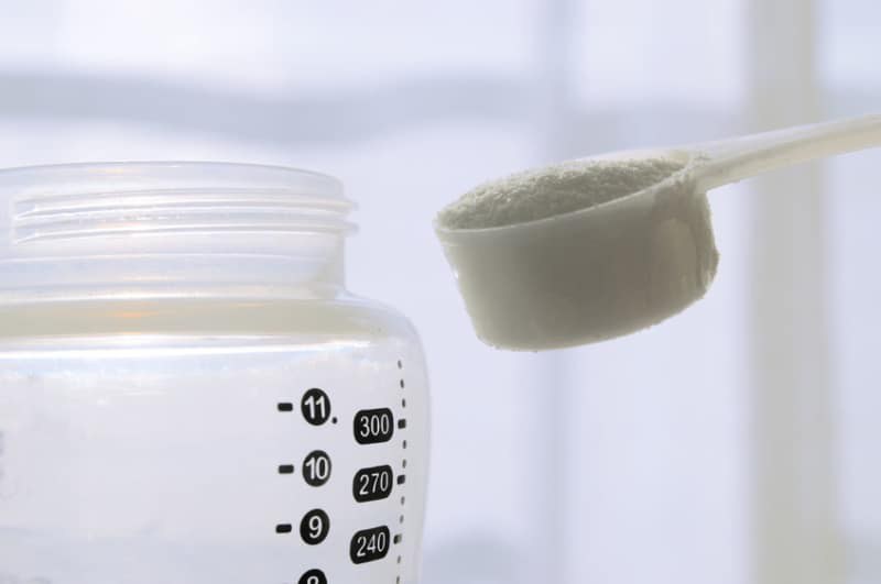 A close-up of a baby bottle with measurement markings and a filled scoop of powdered formula above the bottle opening, ready to be poured in.