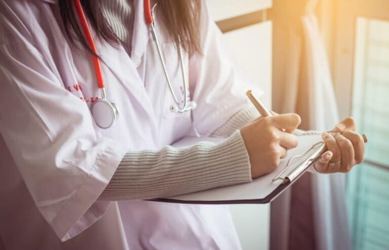 A person wearing a white medical coat and stethoscope writes on a clipboard, with sunlight streaming in from a window.