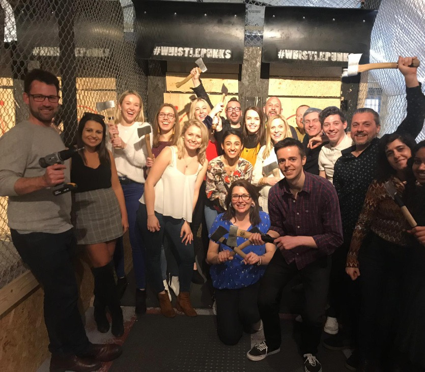 A group of people pose and smile at an indoor axe-throwing venue, holding axes and standing in front of wooden targets with the signs #WHISTLEPUNKS displayed above.