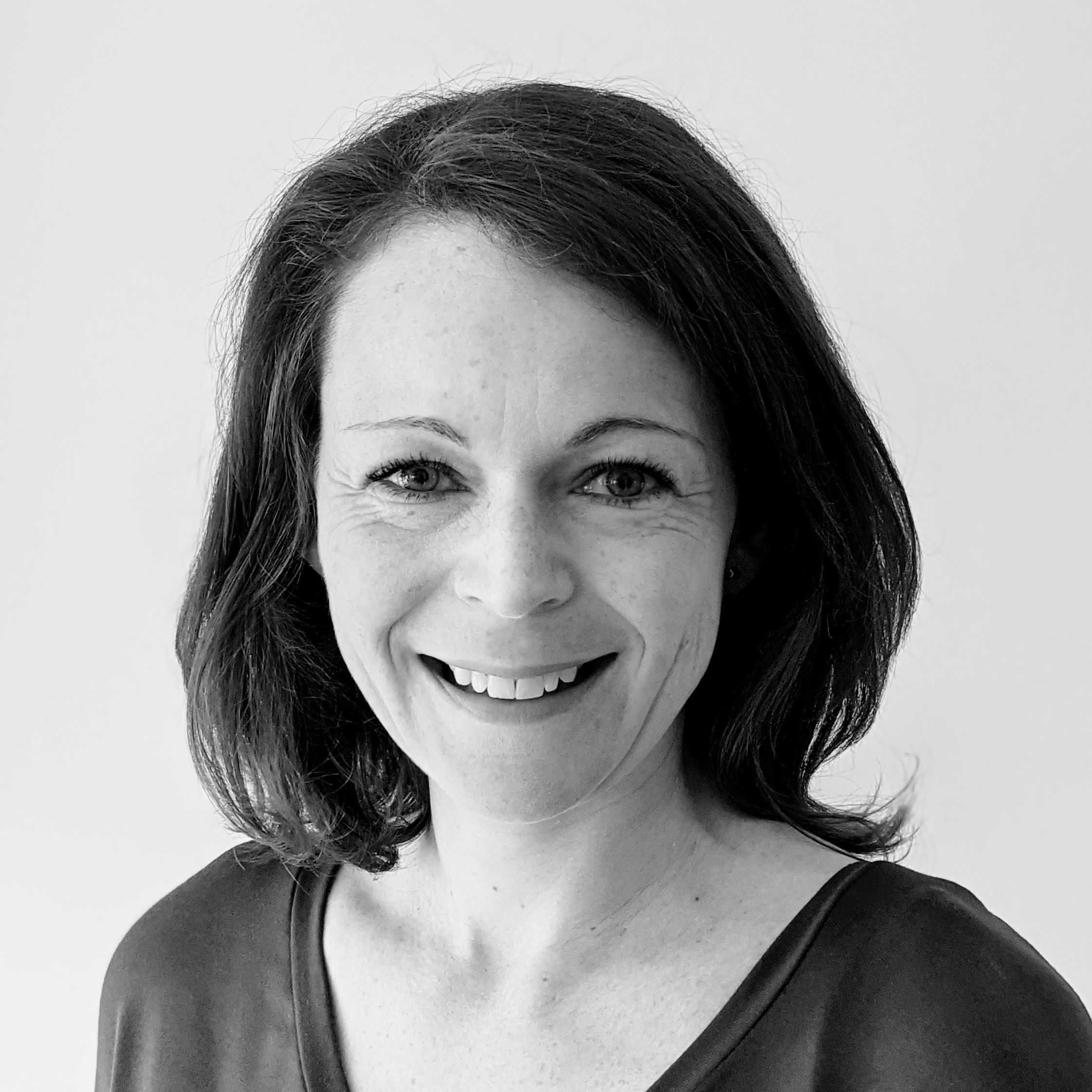 Smiling woman with shoulder-length dark hair, wearing a dark top, poses in front of a plain light background. The photo is in black and white.