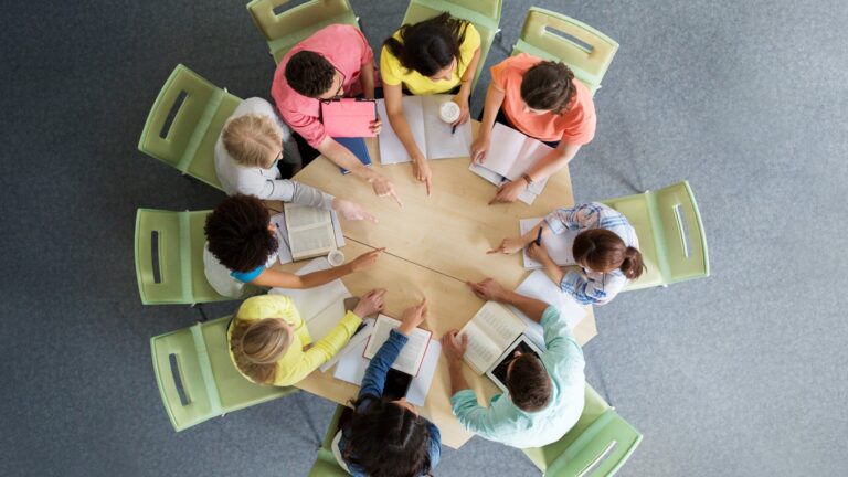 Eight people sit around a round table, studying books and taking notes. The photo is taken from above, showing their books, notebooks, coffee, and colourful clothing. Green chairs surround the table on a grey floor.