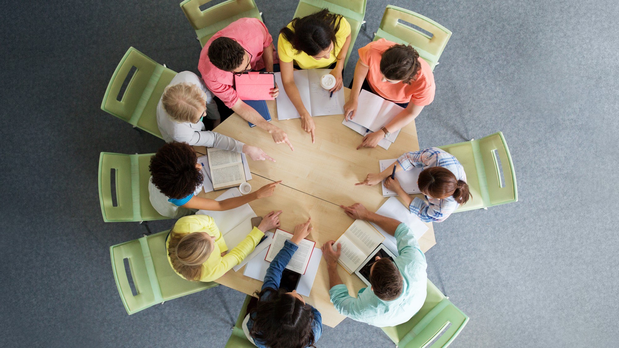 Eight people sit around a round table, studying books and taking notes. The photo is taken from above, showing their books, notebooks, coffee, and colourful clothing. Green chairs surround the table on a grey floor.