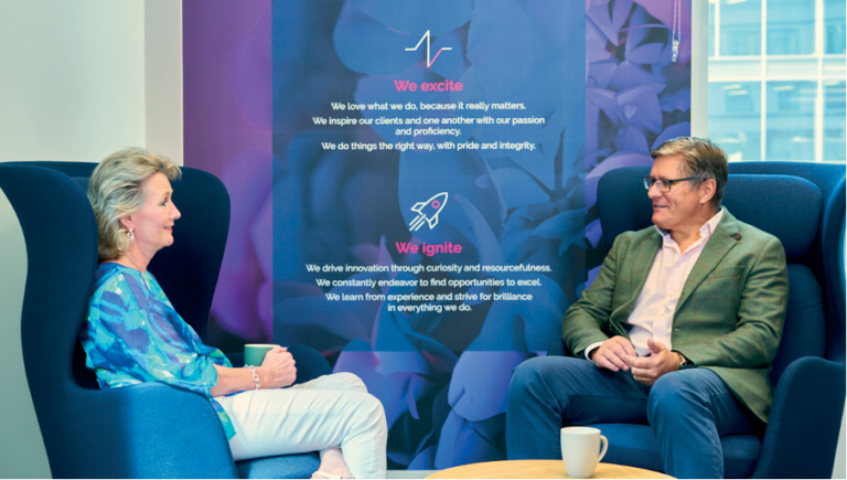 Two people sit in armchairs, conversing and smiling, with coffee cups on the table. Behind them, a purple banner with motivational company values and icons is displayed. A window shows a city view in the background.