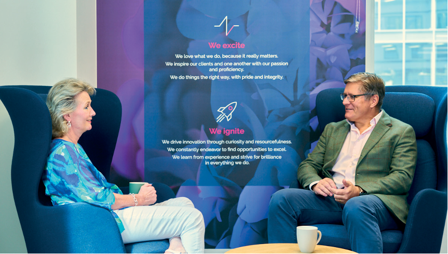 Two people sit in armchairs, conversing and smiling, with coffee cups on the table. Behind them, a purple banner with motivational company values and icons is displayed. A window shows a city view in the background.