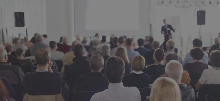 A group of people sit facing a speaker giving a presentation in a bright, modern room with a large projection screen at the front.