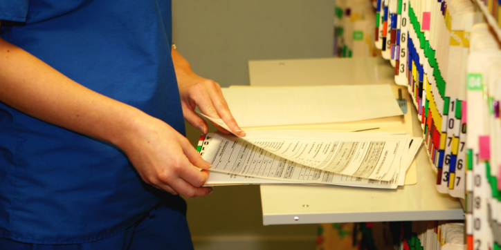 A person in blue scrubs leafs through patient files next to a wall of organised medical records in an office or clinic setting.