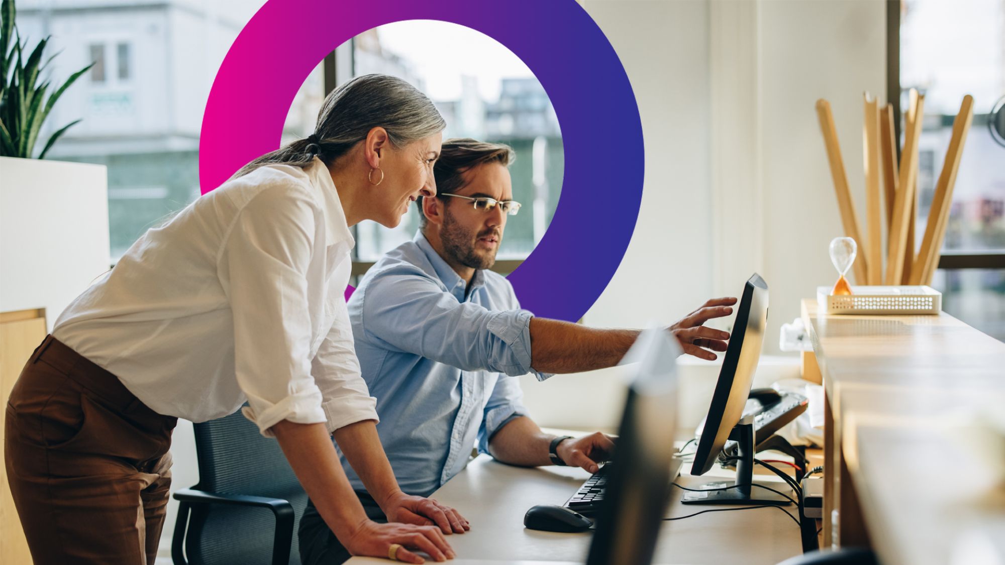 Two people work together at a computer in a modern office. The man points at the screen whilst the woman stands beside him, both focused. A large purple and pink circle decorates the background wall.