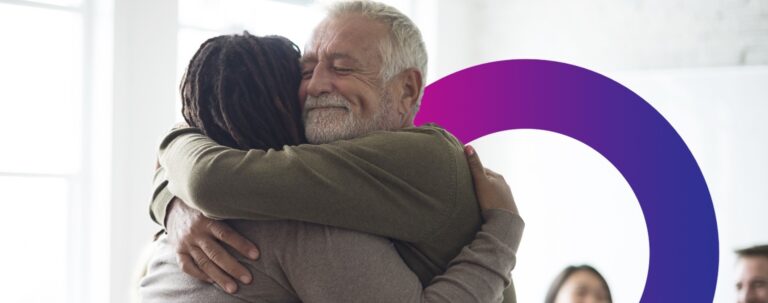 Two people warmly hug each other and smile with eyes closed, expressing comfort and support. Others are visible in the softly lit background, with a purple semi-circle graphic element present.