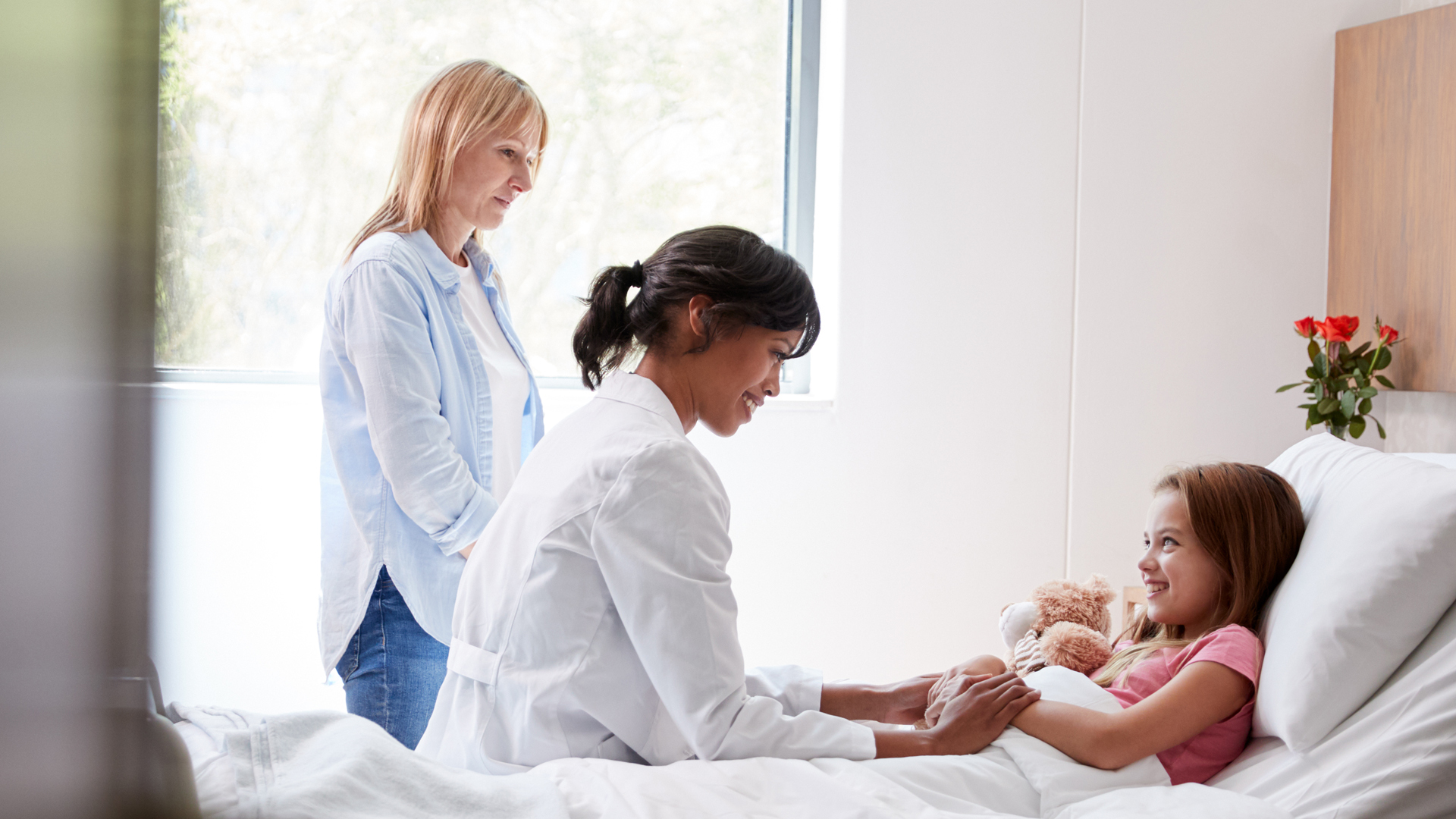 A doctor holds a young girl's hands as she lies in a hospital bed, smiling. A woman stands nearby, watching them. A soft toy and flowers are on the bed.