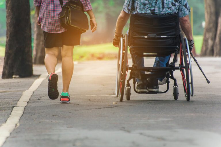 A person in a wheelchair and another person walking beside them travel along a paved park path surrounded by trees, with sunlight filtering through.