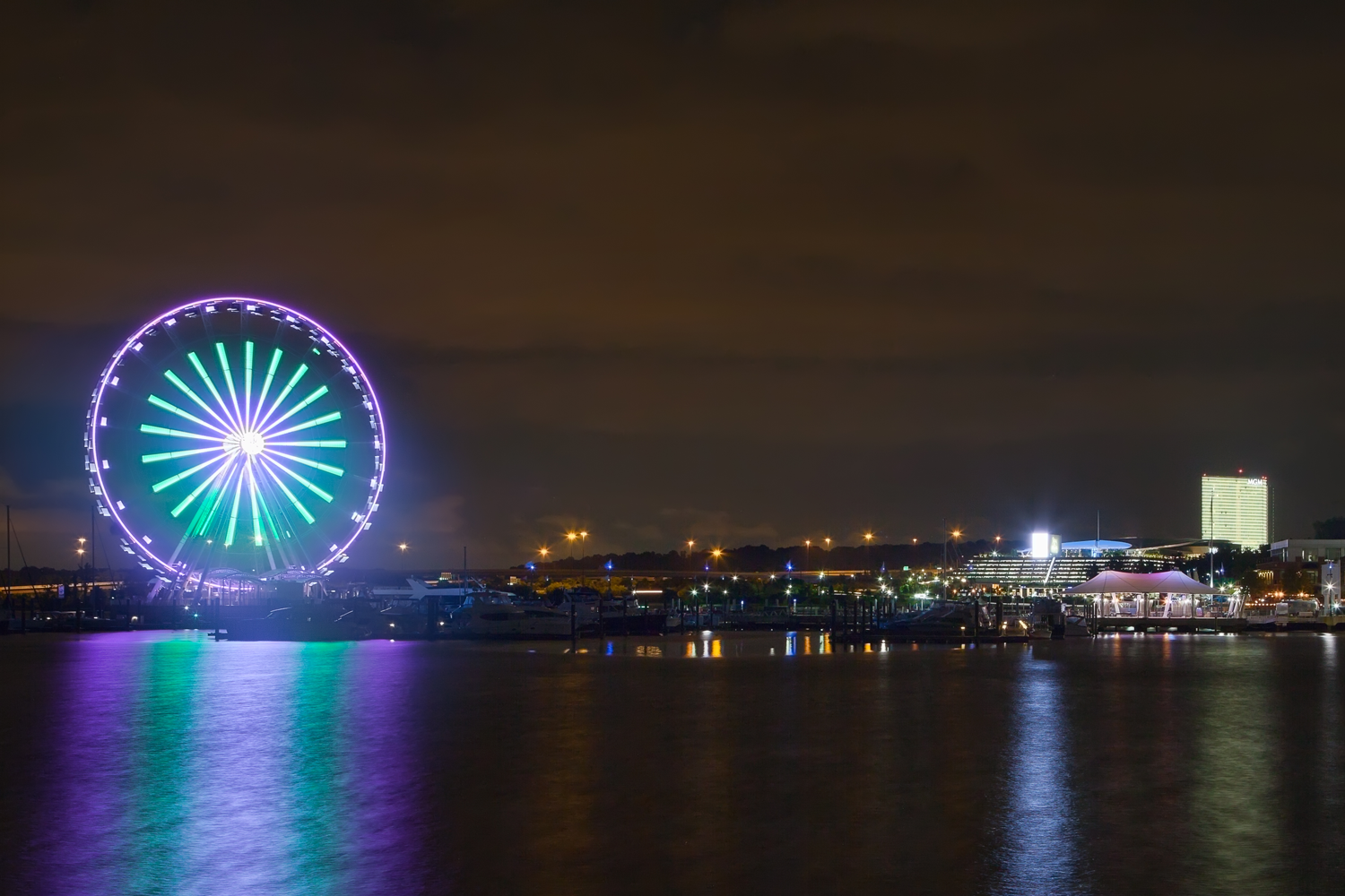 A large, brightly lit Ferris wheel glows with green and purple lights at night, reflecting on the water. City buildings and lights are visible in the background under a cloudy sky.