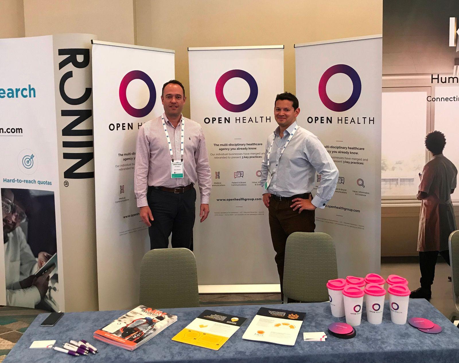 Two men stand in front of three Open Health banners at a conference stand. A table in front of them displays leaflets, magazines, pens, and multiple pink-capped cups. Other stands and displays are visible in the background.
