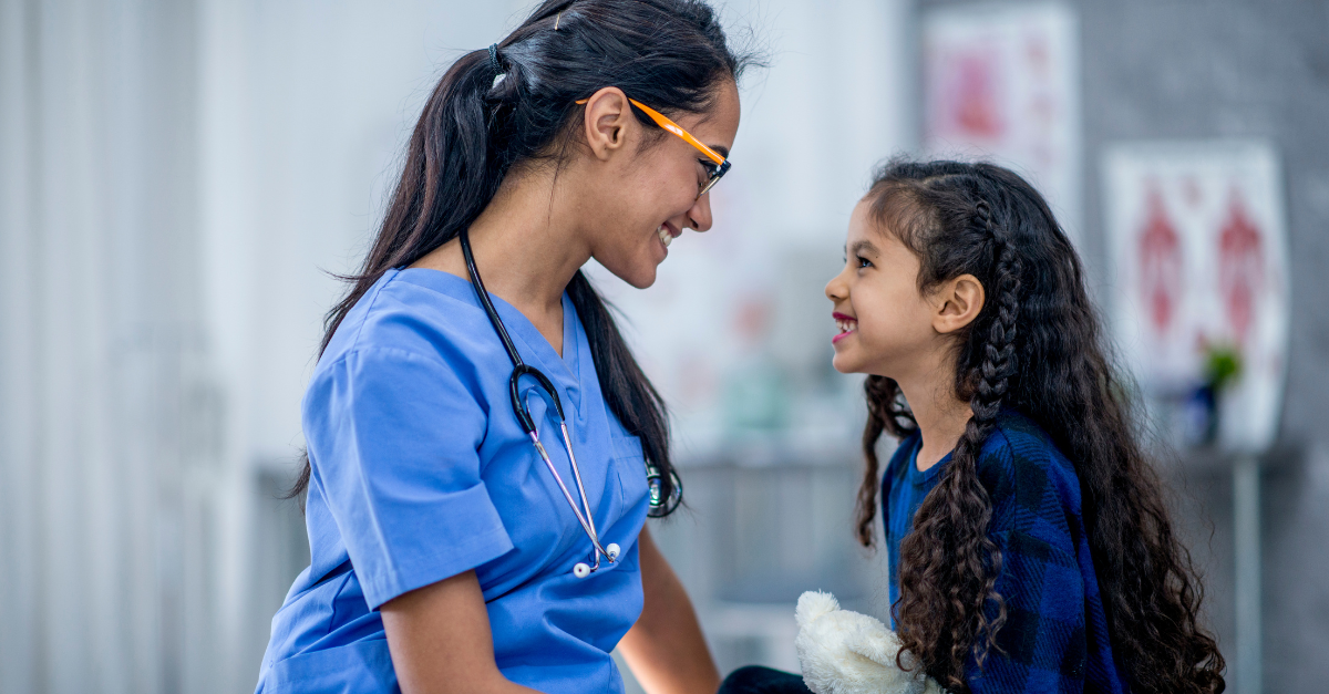 A nurse in blue scrubs smiles at a young girl holding a soft toy. Both are making eye contact and smiling warmly in a medical surgery setting.