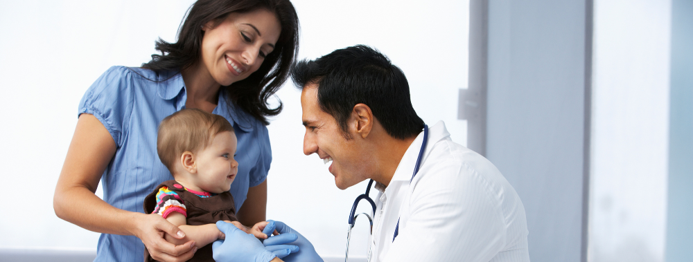 A doctor in a white coat and gloves smiles at a baby whilst giving a vaccine jab, as the baby sits on a woman's lap and looks happy. The woman, likely the mother, smiles at the baby in a bright, modern clinic.