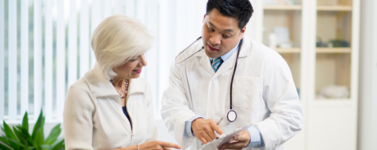 A doctor holding a clipboard talks with an older woman, explaining information to her in a bright medical surgery.