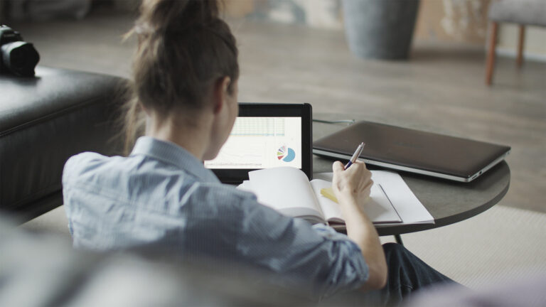 A person sits at a table, writing in a notebook while looking at charts on a tablet. A closed laptop and camera are nearby.