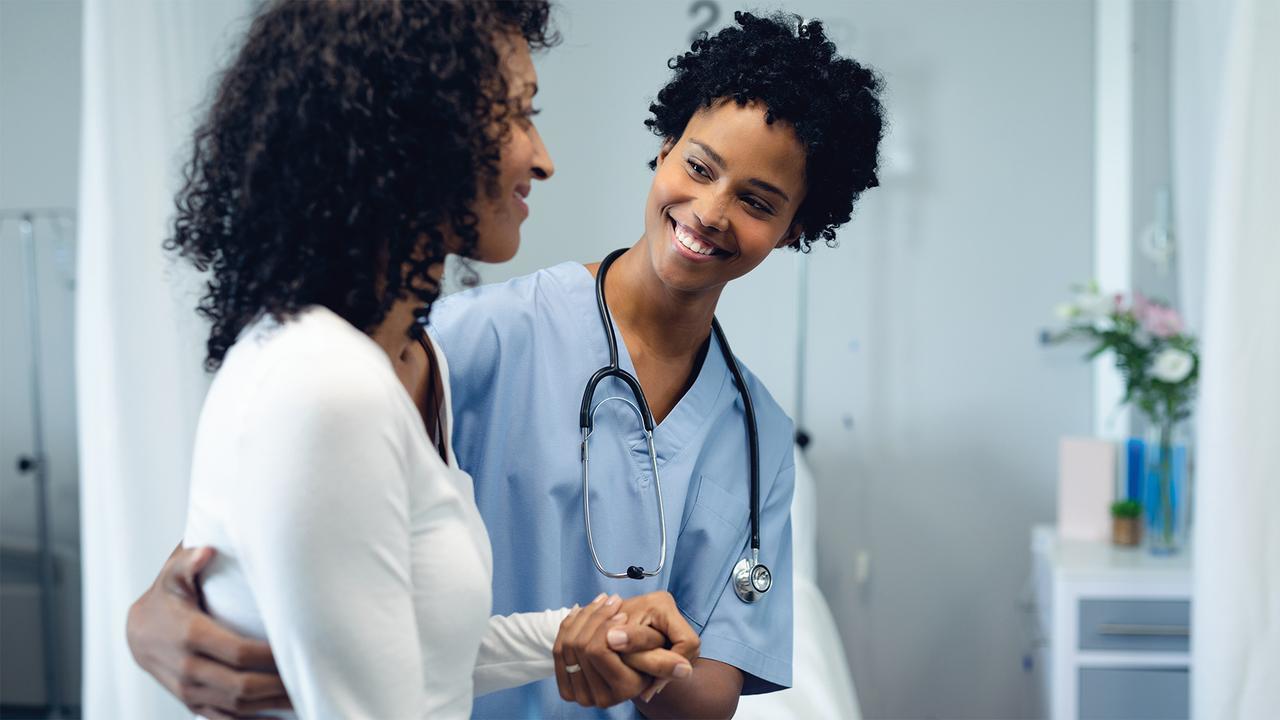 A smiling healthcare professional in scrubs with a stethoscope shakes hands and talks with a patient in a clinic room.