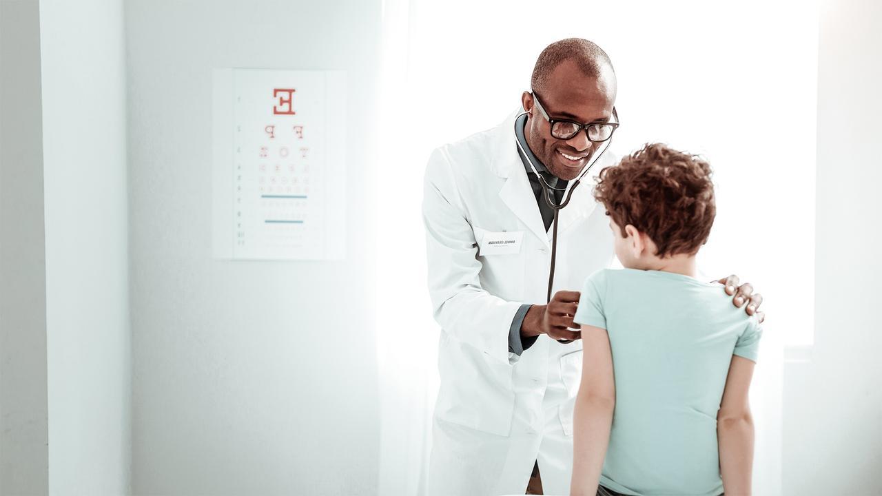 A doctor wearing a white coat and glasses examines a young boy in a light blue shirt with a stethoscope in a brightly lit medical surgery. An eye chart is visible on the wall in the background.
