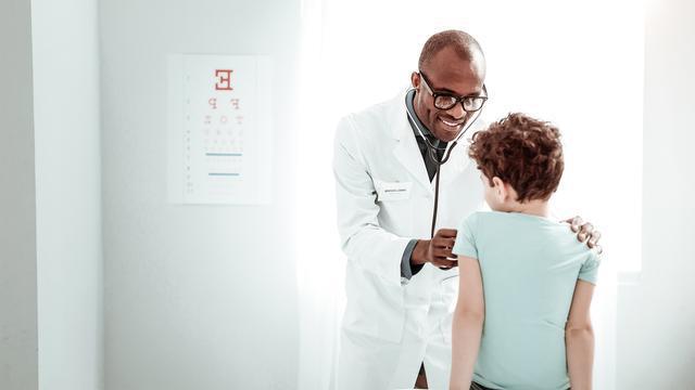 A doctor smiles while using a stethoscope to examine a young boy sitting on an examination table in a brightly lit medical surgery. An eye chart is visible on the wall in the background.
