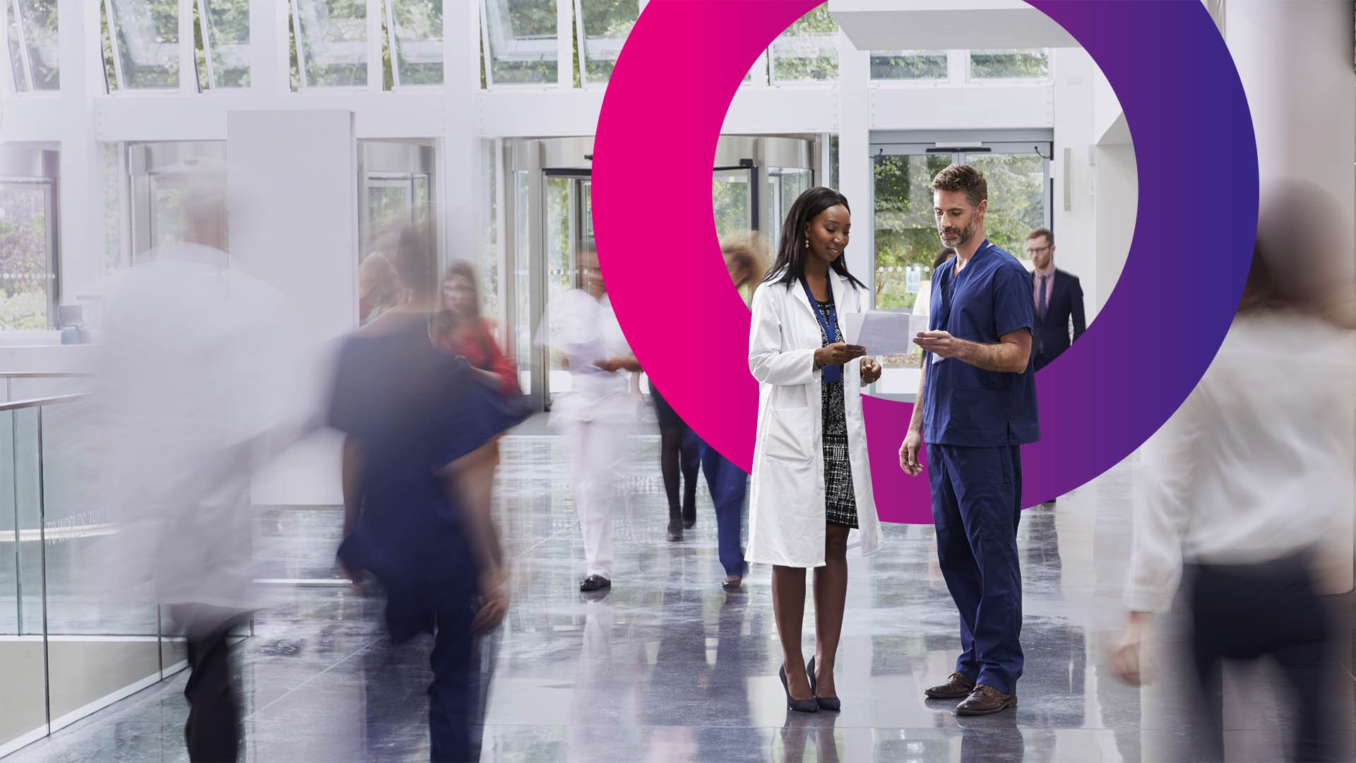 Two healthcare professionals stand and talk in a bright, modern hospital corridor as other people walk by. A large purple and pink circular graphic is behind them.