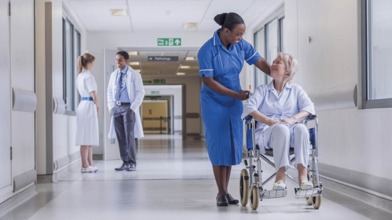 A nurse smiles at an elderly woman in a wheelchair in a hospital corridor. Two doctors stand and talk in the background. The scene is bright and friendly.