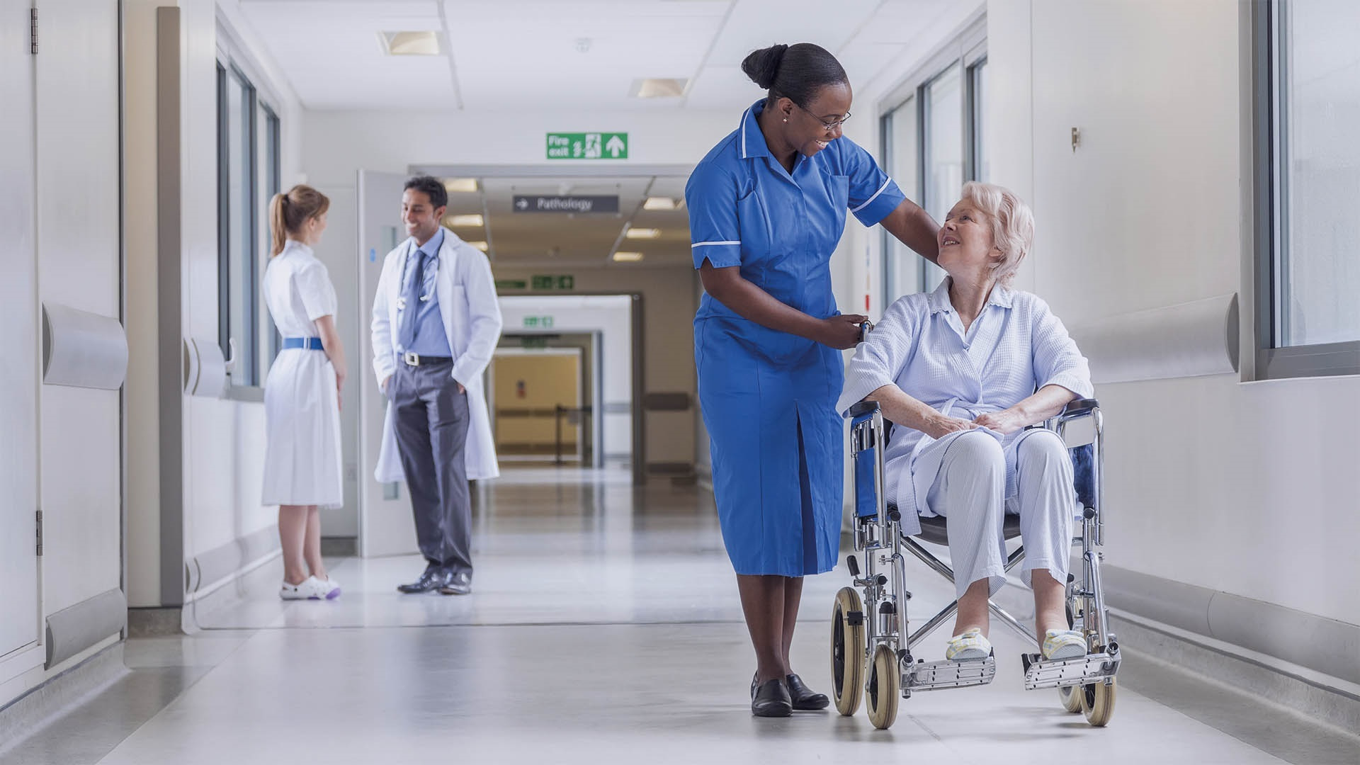 A nurse smiles at an elderly woman in a wheelchair in a hospital corridor. Two doctors stand and talk in the background. The scene is bright and friendly.