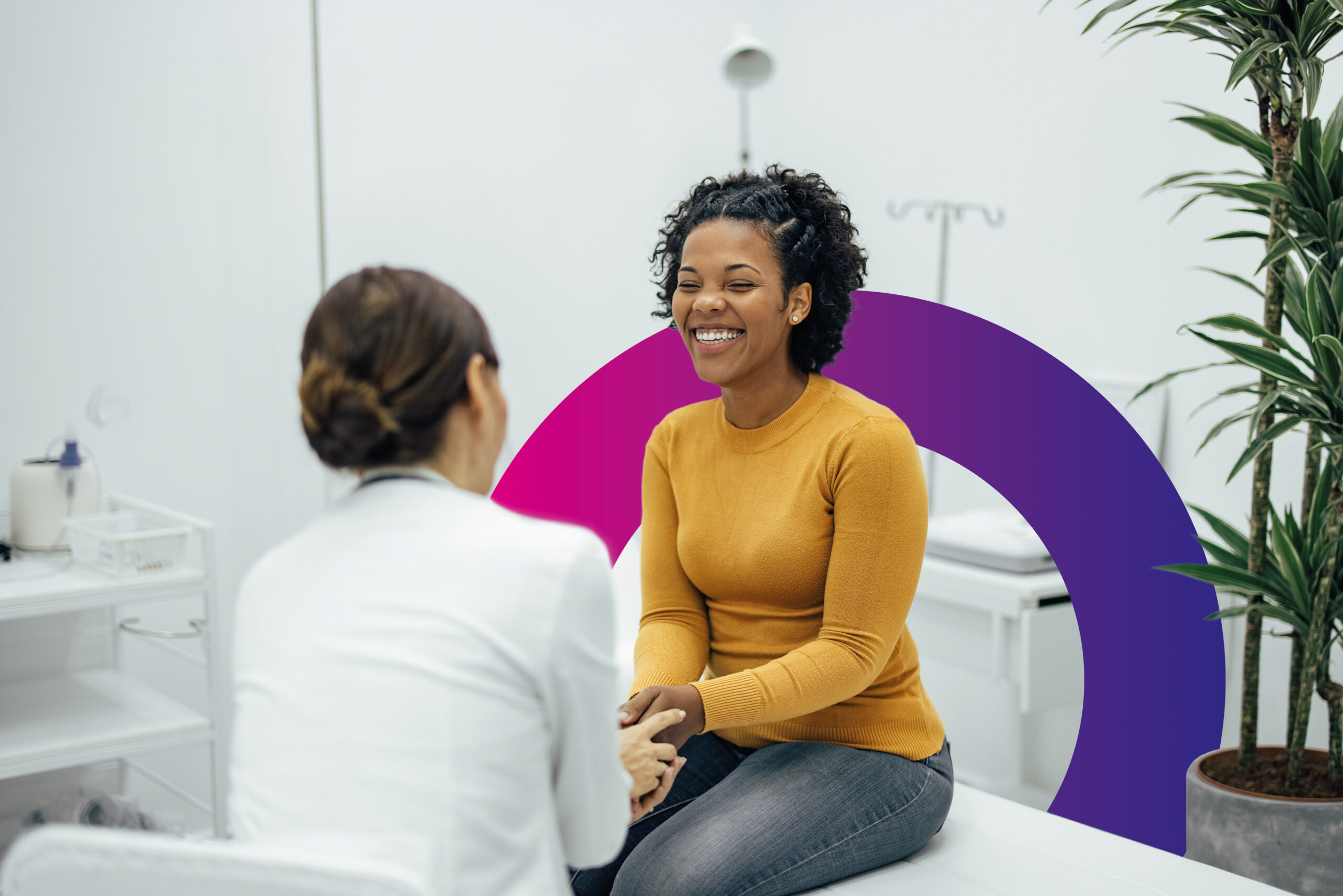 A woman in a mustard jumper smiles whilst talking to a healthcare professional in a white coat in a bright, modern medical surgery with plants and medical equipment in the background.