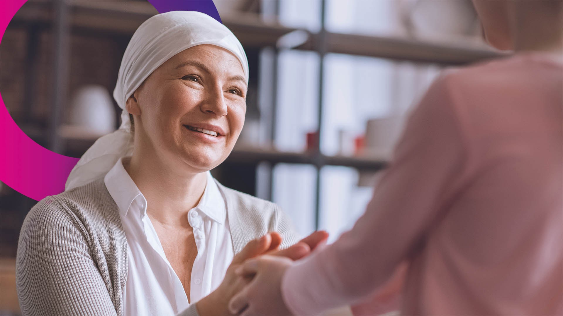 A woman wearing a headscarf smiles warmly whilst holding hands with another person whose back is to the camera.