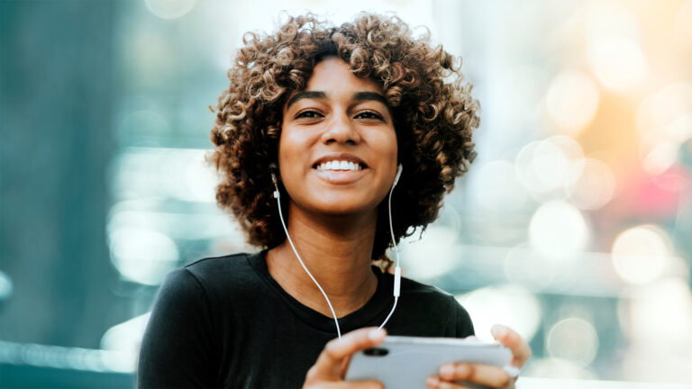 A smiling person with curly hair listens to headphones and holds a mobile phone outdoors with blurred lights in the background.