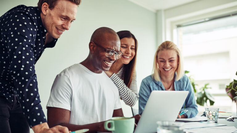 Four people smiling and gathered around a laptop, appearing to collaborate and enjoy themselves in a bright, casual office setting.