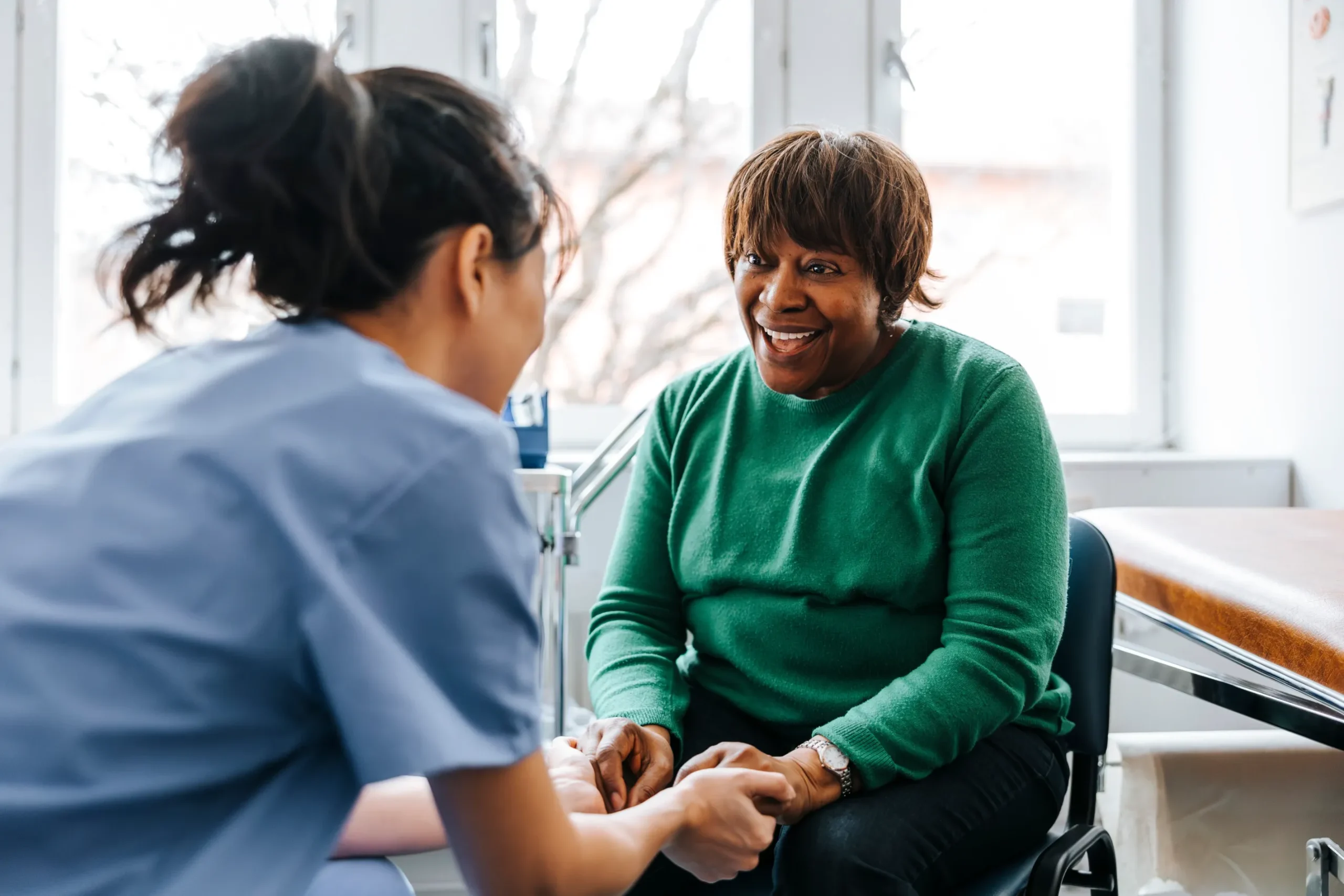 A healthcare worker in scrubs holds the hands of a smiling woman in a green jumper during a conversation in a bright medical surgery.