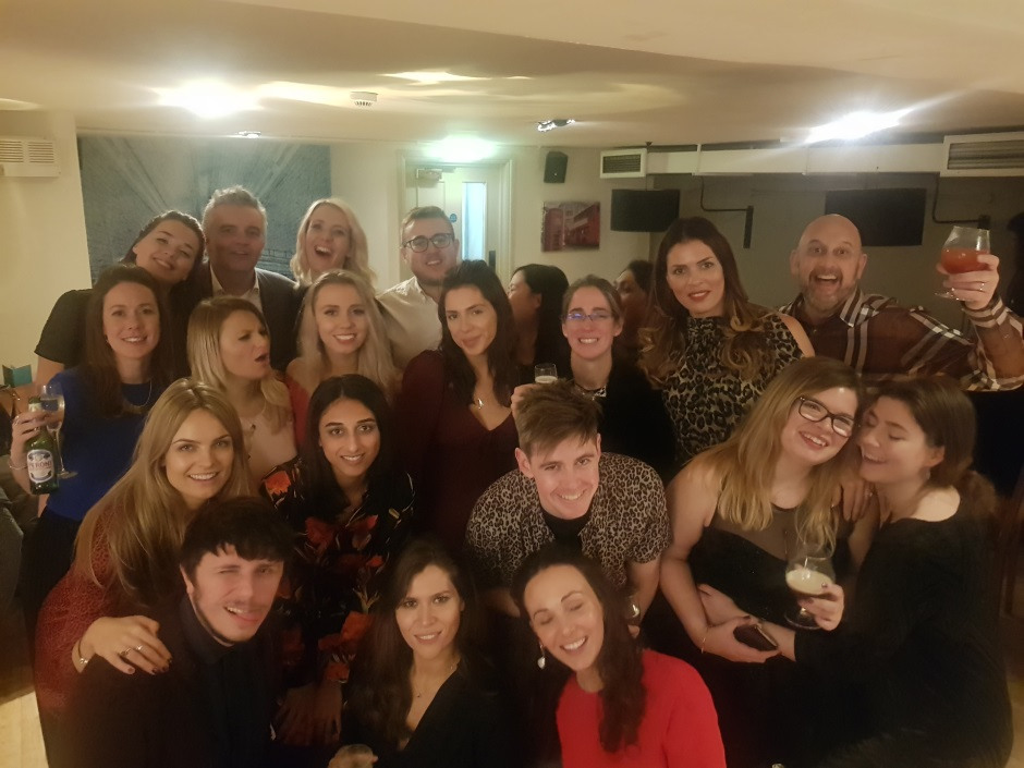A group of people posing together at a party indoors, many smiling and holding drinks. The atmosphere is lively and friendly, with warm lighting and casual attire.
