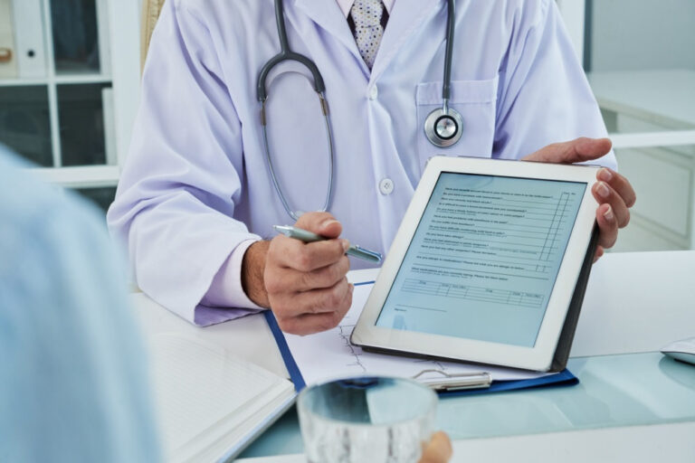 A doctor in a white coat shows a digital form on a tablet to a patient, holding a pen and gesturing towards the screen during a consultation in an office.
