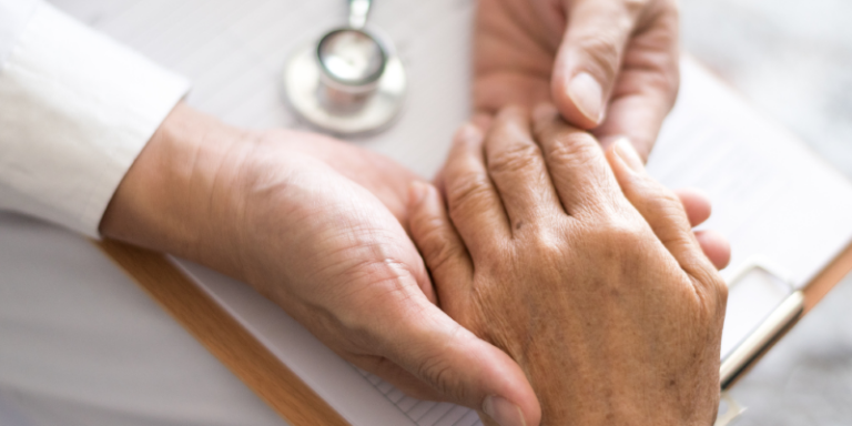 A doctor gently holds a patient's hand in a comforting gesture, with a stethoscope and clipboard visible in the background.