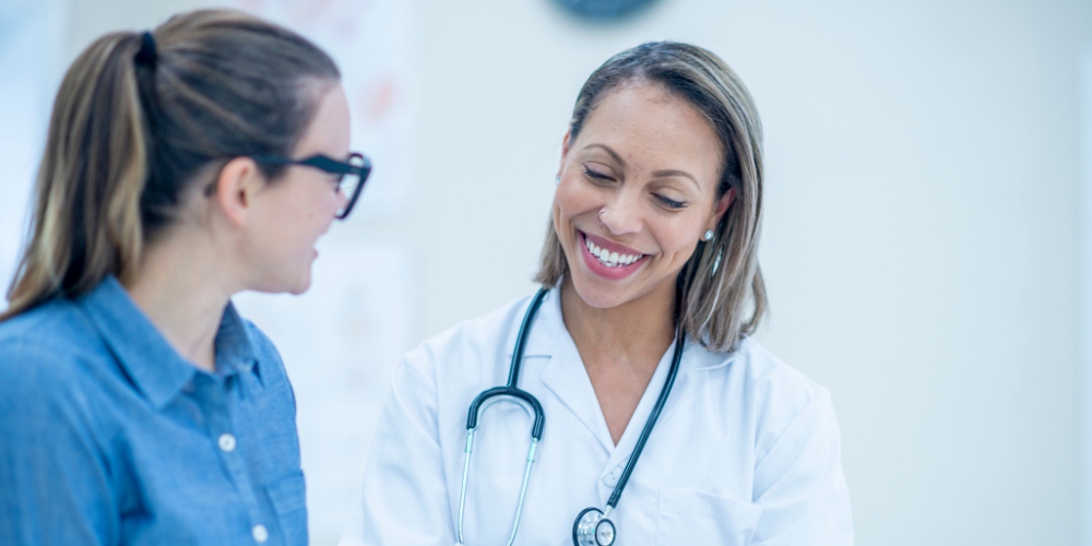 A doctor with a stethoscope smiles whilst talking to a patient wearing glasses. Both women appear to be having a friendly conversation in a medical surgery.