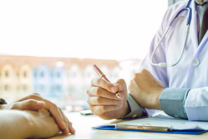 A doctor in a white coat holds a pen and discusses something with a patient, whose hands are visible on the table. A clipboard with papers is in front of the doctor.