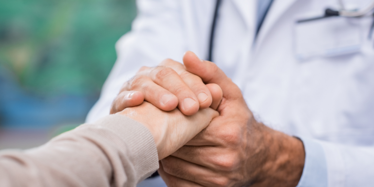 A doctor gently holds a patient's hand in a comforting gesture, both wearing long sleeves. The doctor's white coat and stethoscope are partially visible. The background is softly blurred.