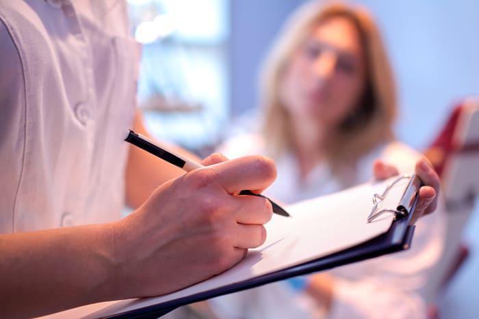 A person writes on a clipboard with a pen, whilst another person sits in the background, slightly out of focus.