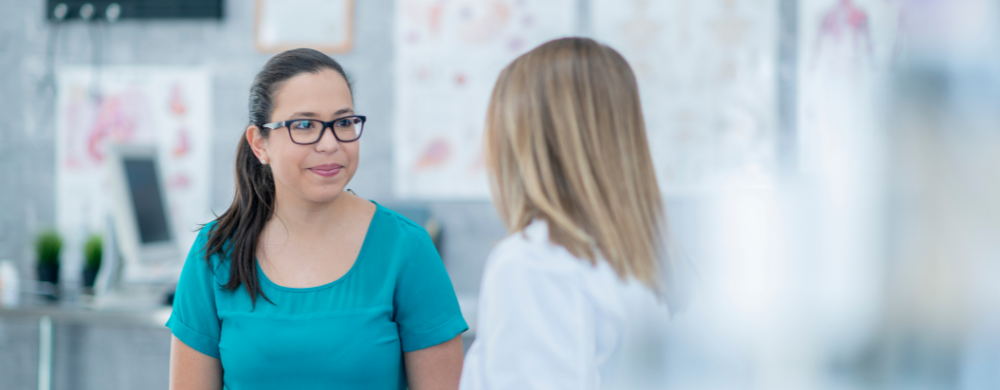 A woman wearing glasses and a teal shirt smiles whilst talking to another woman with blonde hair in a white coat in a bright office or clinic setting.