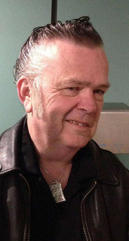 Smiling older man with slicked-back hair, wearing a black leather jacket, black shirt, and a silver necklace, standing in front of a light green background.