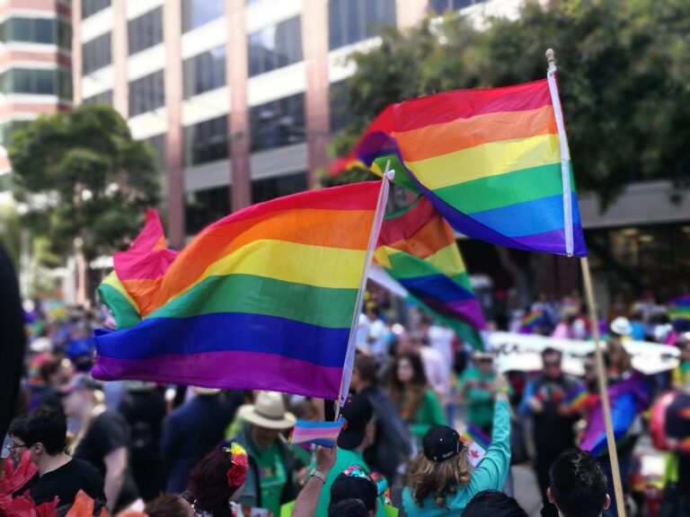 People hold and wave rainbow pride flags at a colourful outdoor event, likely a parade or festival, with a crowd and tall buildings in the background.