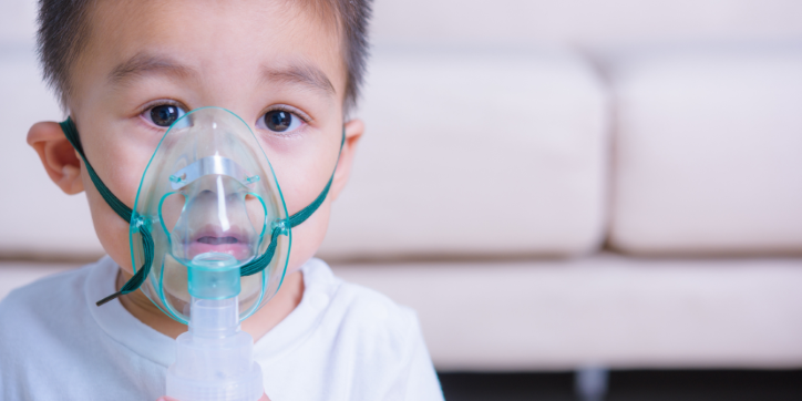 A young child wearing a white shirt uses a nebuliser mask, sitting indoors with a blurred sofa in the background.