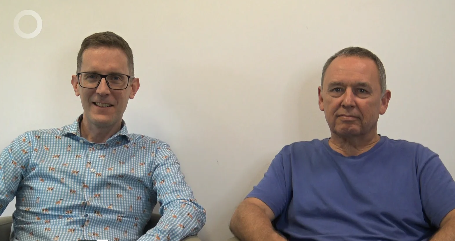 Two men sitting next to each other against a plain light-coloured wall. The man on the left wears glasses and a patterned shirt; the man on the right wears a blue T-shirt.