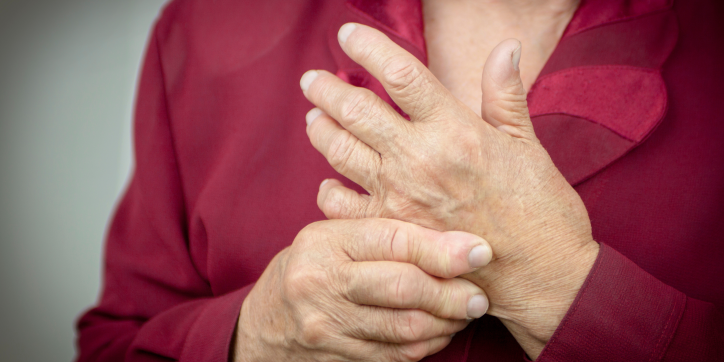 An older person wearing a maroon shirt holds their right wrist with their left hand, suggesting discomfort or pain in the hand or wrist.