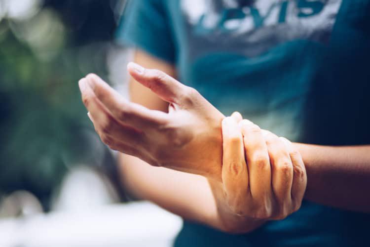 A person in a teal shirt holds their right wrist with their left hand, suggesting wrist pain or discomfort. The background is softly blurred.