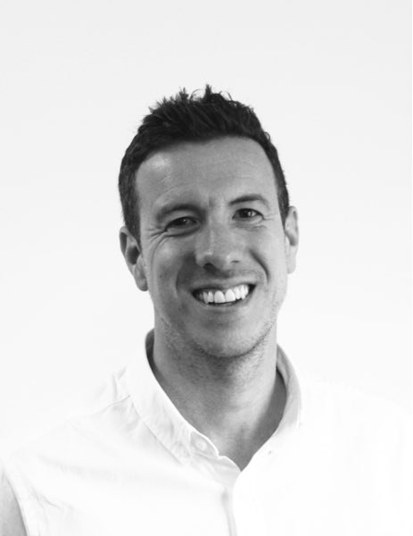 A man with short dark hair smiles at the camera. He is wearing a white, sleeveless collared shirt. The background is plain and light-coloured. The image is in black and white.