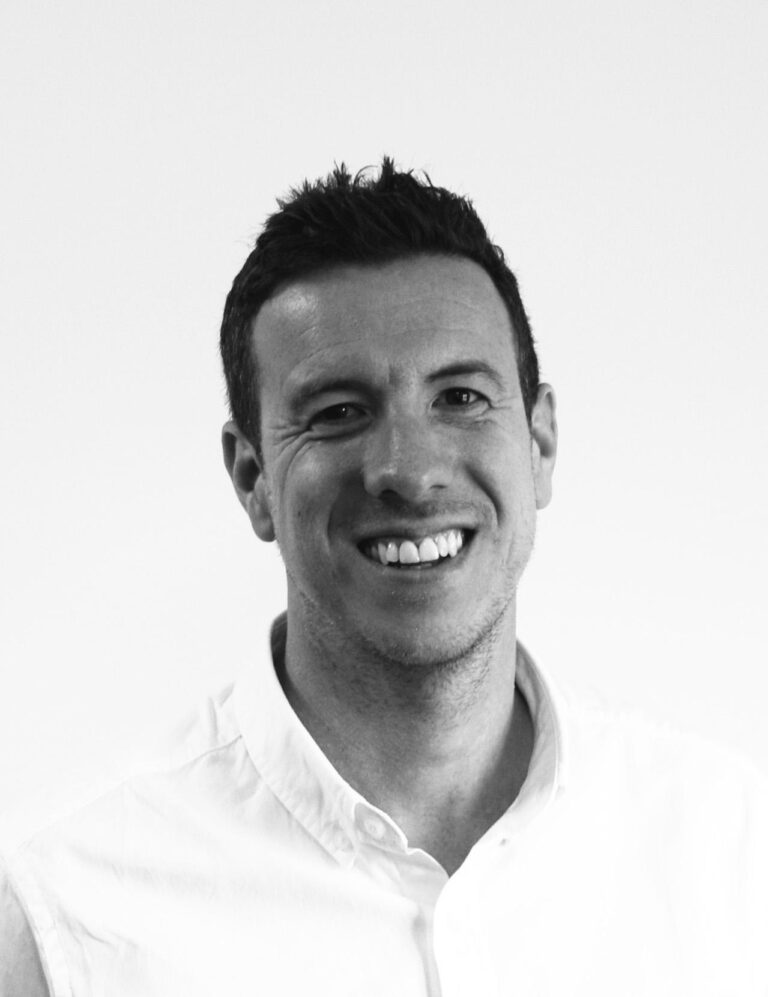 Black and white photo of a man with short dark hair, smiling, wearing a light-coloured button-up shirt, posed against a plain background.