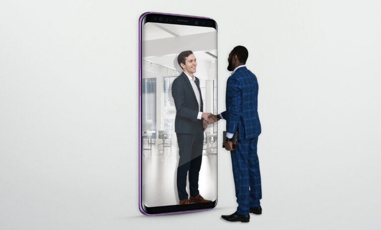 A man in a blue suit shakes hands with another man who appears to be inside a giant mobile phone screen, symbolising virtual or remote business interaction.