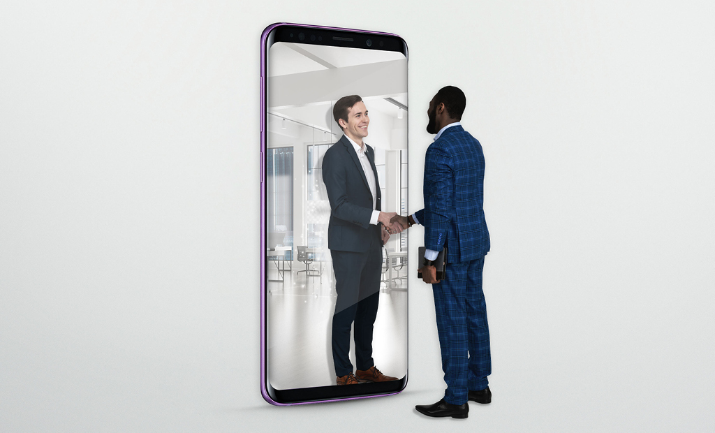 A man in a blue suit shakes hands with another man who appears to be inside a giant mobile phone screen, symbolising virtual or remote business interaction.